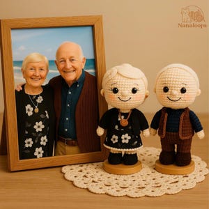 K&ouml;nnte beinhalten: Ein gerahmtes Foto eines &auml;lteren Ehepaares steht neben zwei geh&auml;kelten Puppen. Die Puppen tragen Miniatur-Outfits, und die Puppen-Frau tr&auml;gt ein schwarzes Kleid mit wei&szlig;en Blumenmustern.