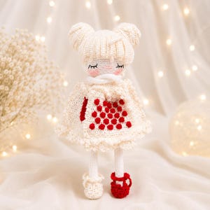 May include: A handmade crochet doll in a cream-colored dress with red accents. The doll has a white hat with two buns, closed eyes, and red shoes. The dress features a red heart design.