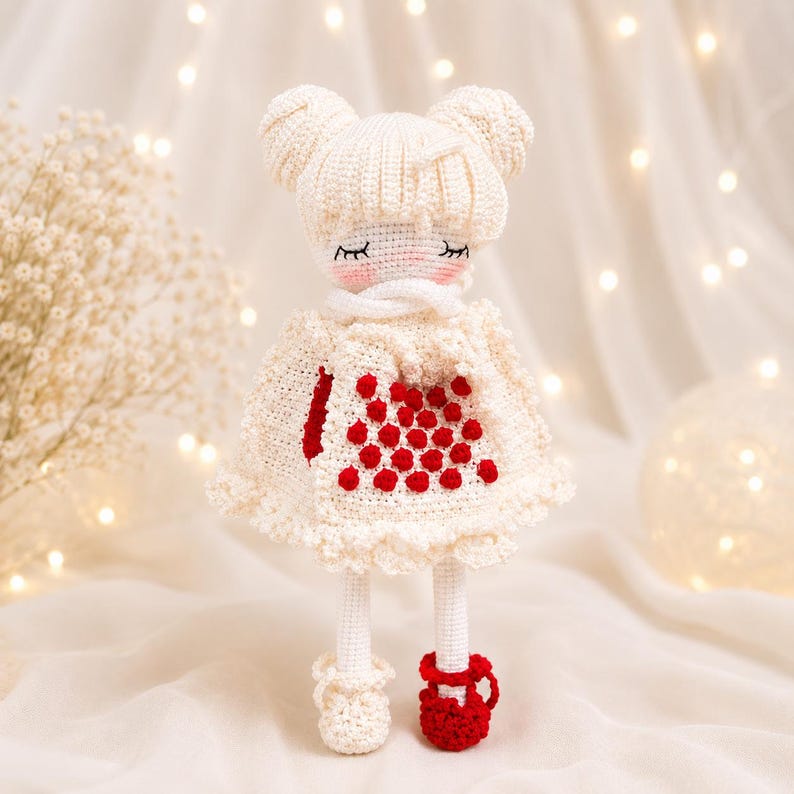 May include: A handmade crochet doll in a cream-colored dress with red accents. The doll has a white hat with two buns, closed eyes, and red shoes. The dress features a red heart design.