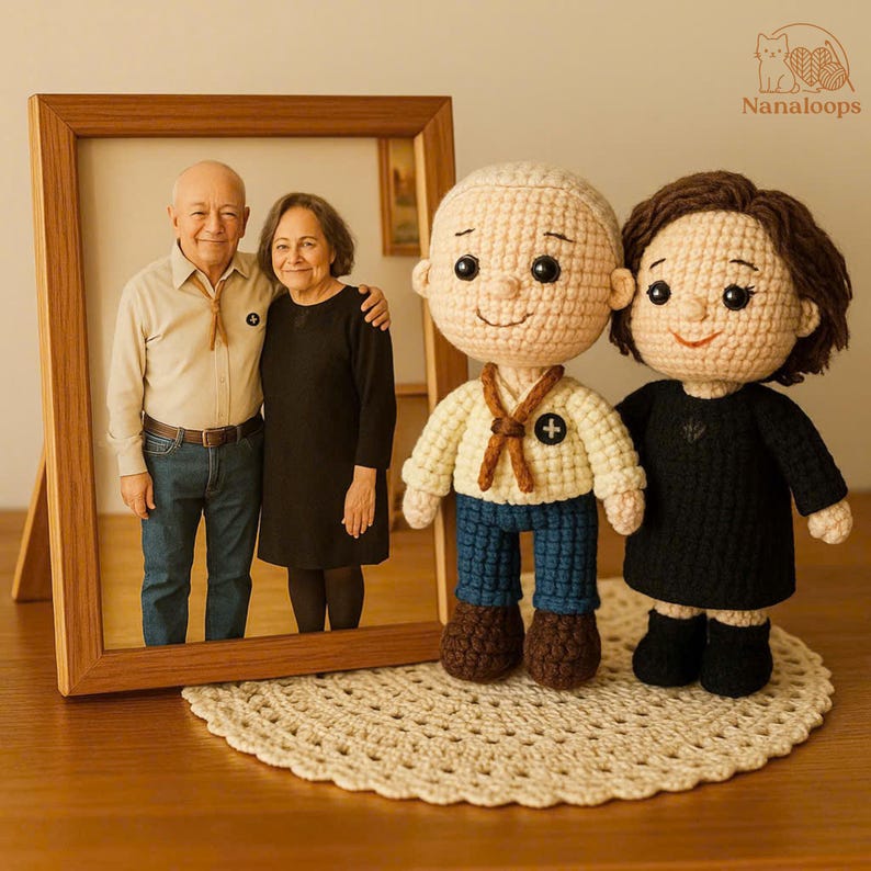 K&ouml;nnte beinhalten: Ein gerahmtes Foto eines &auml;lteren Ehepaares steht neben zwei geh&auml;kelten Puppen, einem Mann und einer Frau. Die Puppen sind beige mit dunklem Haar und tragen ein cremefarbenes Hemd, blaue Hosen und ein schwarzes Kleid. Die Puppen stehen auf einer geh&auml;kelten Tischdecke.
