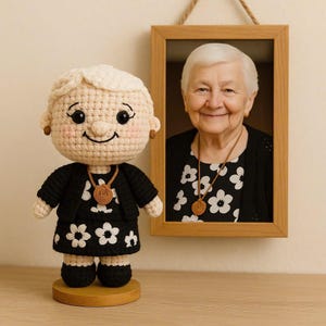 K&ouml;nnte beinhalten: Geh&auml;kelte Puppe einer &auml;lteren Frau in einem schwarzen Outfit mit wei&szlig;en Blumenmustern, einer Halskette und Ohrringen. Im Hintergrund h&auml;ngt ein gerahmtes Foto einer l&auml;chelnden &auml;lteren Frau. Die Puppe steht auf einem Holzsockel.
