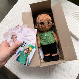 K&ouml;nnte beinhalten: Handgefertigte H&auml;kelpuppe mit Brille, gr&uuml;nem Oberteil und schwarzer Hose in einem Karton. Die Puppe hat braunes Haar und tr&auml;gt eine Brille. Karten mit dem Text "Danke" und "Wie man sich k&uuml;mmert" sind enthalten.