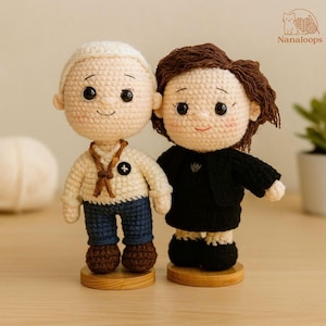 K&ouml;nnte beinhalten: Zwei handgefertigte H&auml;kelpuppen, eine mit einem cremefarbenen Pullover, blauen Jeans und braunen Schuhen, die andere in einem schwarzen Outfit. Beide Puppen haben l&auml;chelnde Gesichter, dunkle Augen und sind auf Holzsockeln montiert.