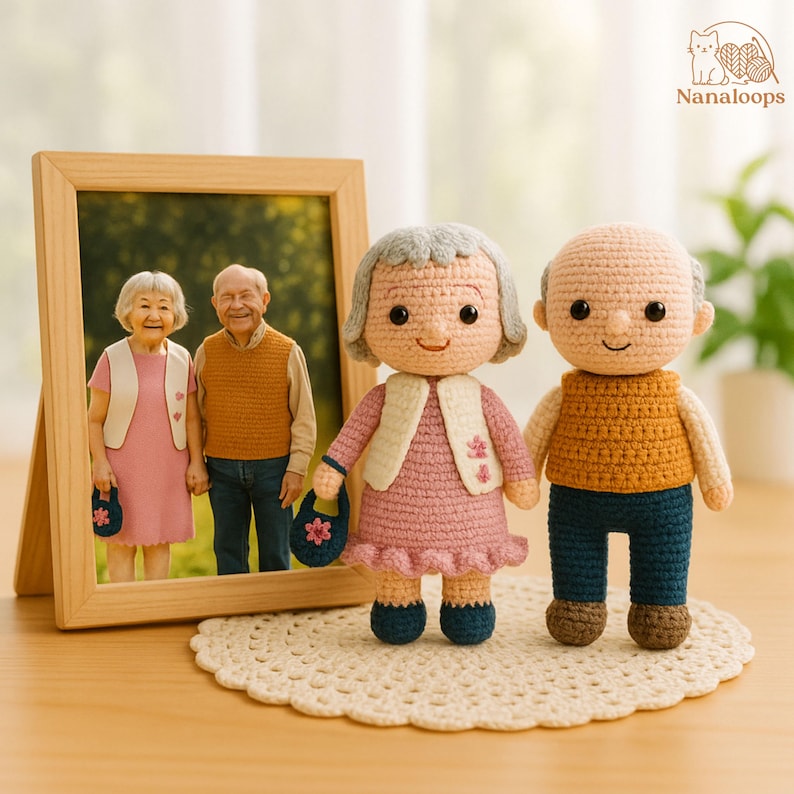 K&ouml;nnte beinhalten: Handgefertigte H&auml;kelpuppen eines &auml;lteren Ehepaares, die neben einem gerahmten Foto eines &auml;lteren Ehepaares stehen. Die Puppen tragen rosa, wei&szlig;e, blaue und braune Kleidung. Die Puppen stehen auf einer wei&szlig;en geh&auml;kelten Tischdecke.