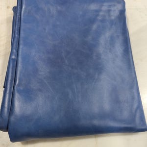 Puede incluir: Una pieza de cuero azul doblada. El cuero tiene una superficie lisa, ligeramente texturizada, con variaciones de color, creando una apariencia natural y envejecida. Los bordes del cuero son visibles, mostrando el grosor y la calidad del material.