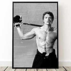 May include: Black and white framed print of a shirtless hockey player holding a hockey stick. The player wears black hockey pants and gloves. The background is a grey ice rink.