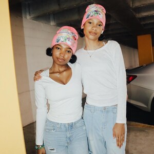 May include: Two people wearing matching pink beanies with colorful cartoon graphics, white long-sleeve shirts, and light blue jeans. They are standing close together, with one person's arm around the other's shoulder.