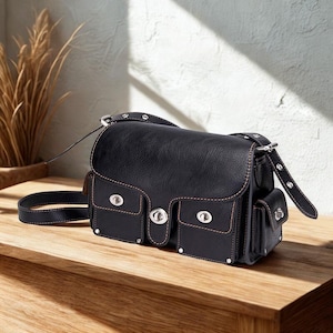 May include: Black satchel bag with silver hardware and multiple pockets. The bag has a long adjustable strap and a flap closure. The stitching is a light brown color, and the bag is sitting on a wooden surface.
