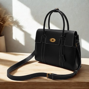 May include: Black leather satchel bag with a top handle and a detachable shoulder strap. The bag features white stitching, a gold-tone clasp, and a flap closure. The bag is sitting on a wooden surface.
