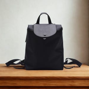 May include: Black backpack with a flap closure and a top handle. The flap is gray and secured with a snap. The backpack has adjustable shoulder straps and a zipper closure. The bag is sitting on a wooden surface.