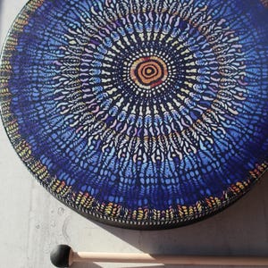 May include: A round drum with a vibrant, intricate blue and purple mandala design. The drum has a black rim and is accompanied by a wooden drumstick with a black head. The design features a central circular pattern with radiating details.