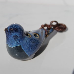 May include: A ceramic bird-shaped ocarina in shades of blue and gray. The musical instrument has three finger holes and a brown cord attached. The bird's beak is accented with gold. The ocarina is a unique musical instrument.