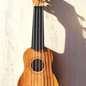 May include: A small ukulele with a light brown wooden body and a black fretboard. The instrument has four white strings and a round sound hole with decorative carvings. The ukulele is approximately 21 inches long.