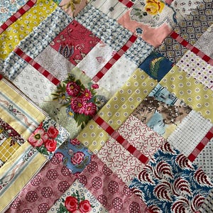 May include: A patchwork quilt constructed from various fabric squares featuring diverse floral, geometric, and striped patterns in shades of red, yellow, blue, pink, and white, all joined together by thin red fabric strips in a grid arrangement.