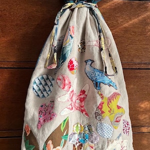 May include: A beige fabric bag with a drawstring closure. The bag is decorated with colorful fabric scraps in a patchwork style, including a blue bird, flowers, and geometric patterns. The text "Petranella Bag Take an Element" is printed at the top of the bag. The text "Margaret Sampson George" is printed at the bottom of the bag.