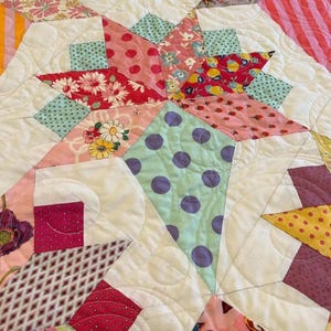 May include: A close-up of a quilt with a patchwork design featuring various floral and polka dot patterns in shades of pink, red, green, yellow, and purple. The quilt is quilted with a white thread and has a white background.