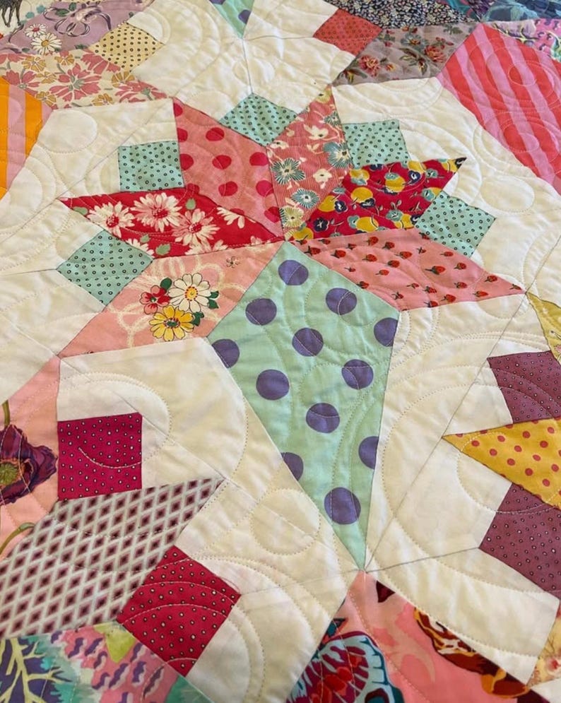 May include: A close-up of a quilt with a patchwork design featuring various floral and polka dot patterns in shades of pink, red, green, yellow, and purple. The quilt is quilted with a white thread and has a white background.