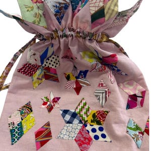 May include: A pink drawstring bag with star-shaped patchwork designs in various colors and patterns. The bag features the text "Seeing Stars" and "Take an Element Margaret Sampson George". The top of the bag has a floral design.