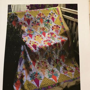 Puede incluir: Una colcha colorida con fondo amarillo y un patrón de estrellas rojas, rosas y blancas. La colcha está doblada y drapeada sobre una barandilla blanca. El título "RUBY ROSEHIPS" y el nombre "Margaret Sampson-George" están impresos sobre la colcha.