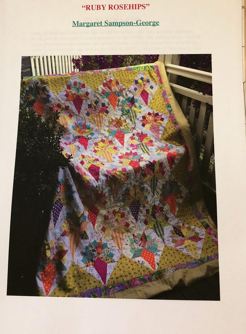May include: A colorful quilt with a yellow background and a pattern of red, pink, and white stars. The quilt is folded and draped over a white railing. The title "RUBY ROSEHIPS" and the name "Margaret Sampson-George" are printed above the quilt.