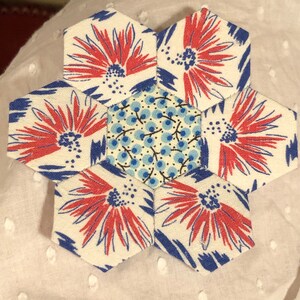 May include: A white fabric hexagon flower block with six red and blue floral patterns and a central hexagon with a blue and white floral pattern.