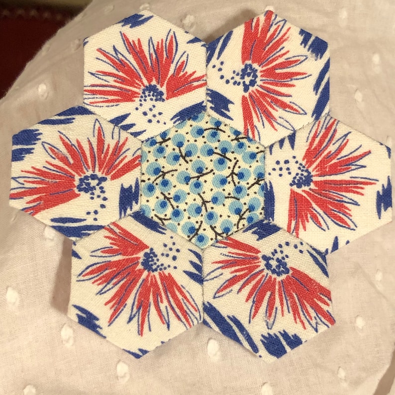 May include: A white fabric hexagon flower block with six red and blue floral patterns and a central hexagon with a blue and white floral pattern.