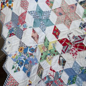 May include: A handmade quilt with a star pattern. The quilt features a variety of colorful floral and geometric patterned fabrics, including red, blue, and yellow, set against white fabric. The quilt is edged with a striped fabric.