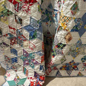May include: A vintage quilt with a geometric pattern. The quilt features a patchwork design with various fabric patterns in red, blue, green, and yellow, set against a white background. The edges are finished with a striped border.