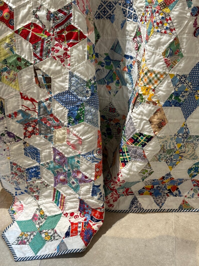 May include: A vintage quilt with a geometric pattern. The quilt features a patchwork design with various fabric patterns in red, blue, green, and yellow, set against a white background. The edges are finished with a striped border.