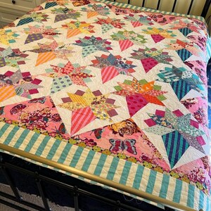 May include: A colorful patchwork quilt with a starburst pattern in shades of pink, blue, green, and yellow. The quilt is made with a variety of fabrics and has a white background. The quilt is displayed on a bed with a black metal frame.