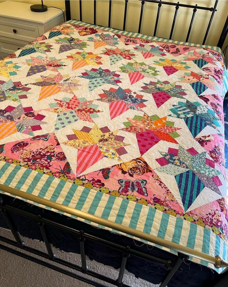 May include: A colorful patchwork quilt with a starburst pattern in shades of pink, blue, green, and yellow. The quilt is made with a variety of fabrics and has a white background. The quilt is displayed on a bed with a black metal frame.