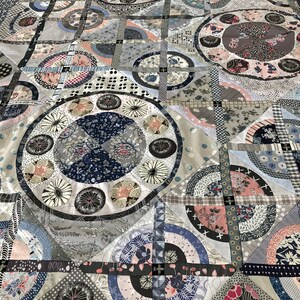 May include: A quilt made of various fabrics in shades of gray, blue, and pink. The quilt features a repeating pattern of circles and squares. The quilt is made of a variety of fabrics, including cotton, linen, and silk.
