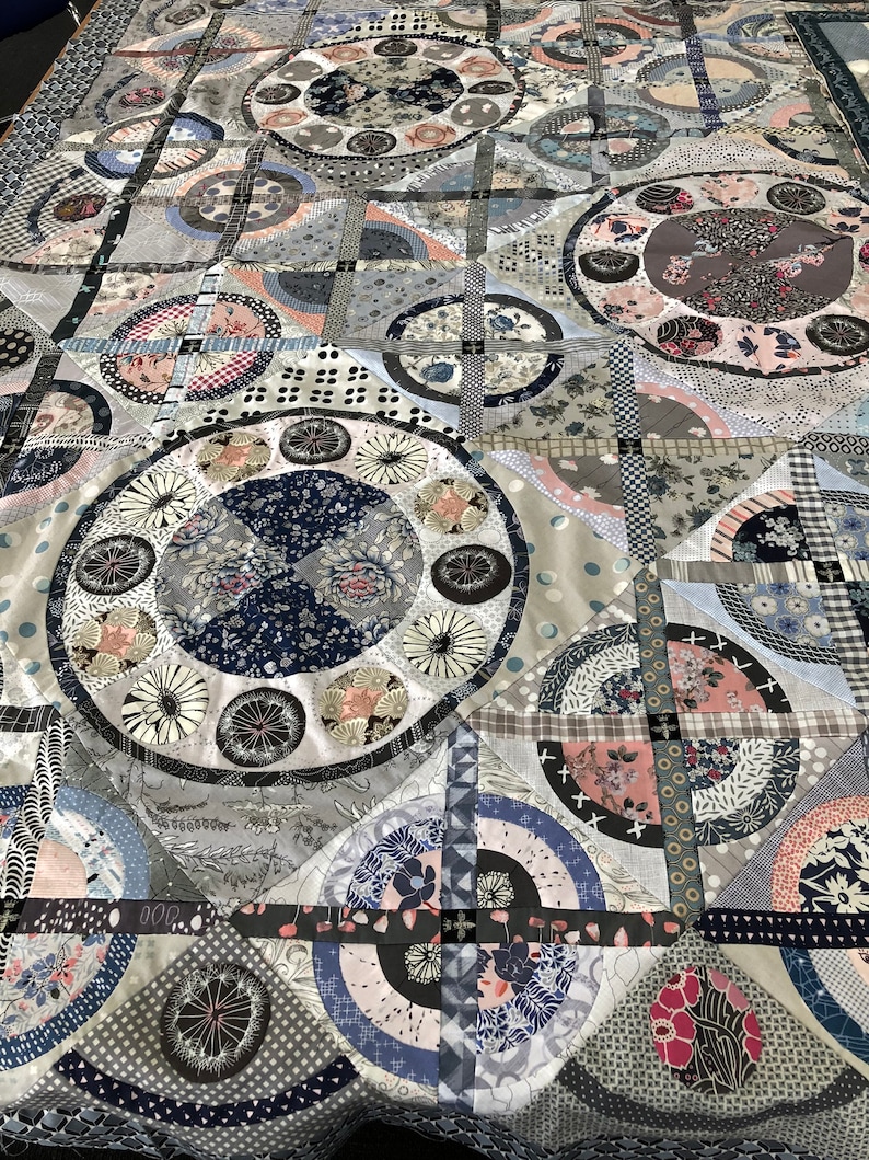 May include: A quilt made of various fabrics in shades of gray, blue, and pink. The quilt features a repeating pattern of circles and squares. The quilt is made of a variety of fabrics, including cotton, linen, and silk.