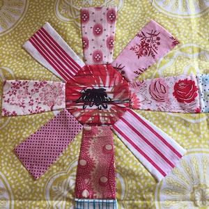 May include: A patchwork quilt block with a starburst design. The block is made of pink and red fabric with various patterns, including stripes, polka dots, and floral prints. The centre of the block is a red fabric with a black design.