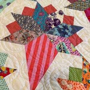 May include: A close-up of a quilt with a star pattern. The quilt is made of various fabrics with floral and geometric designs in shades of pink, red, green, blue, and white. The quilt is quilted with a swirling pattern.