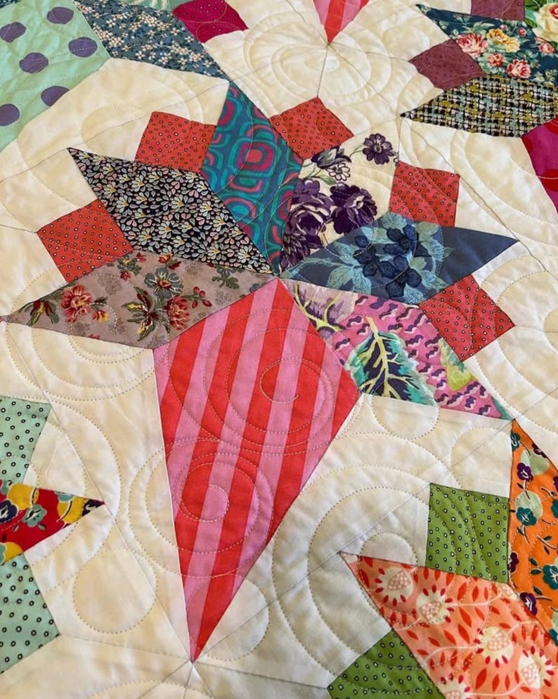 May include: A close-up of a quilt with a star pattern. The quilt is made of various fabrics with floral and geometric designs in shades of pink, red, green, blue, and white. The quilt is quilted with a swirling pattern.