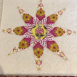 May include: A star-shaped fabric quilt block featuring eight pointed rays made from a white, pink, and olive green ikat-style patterned textile. The center is a yellow hexagon with a red and green floral illustration of a single rose.