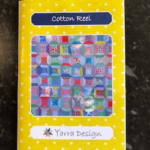 Pode incluir: Um livreto amarelo com estrelas brancas impressas na capa. O livreto é intitulado "Cotton Reel" e apresenta um padrão de colcha colorido com o texto "Yarra Design".
