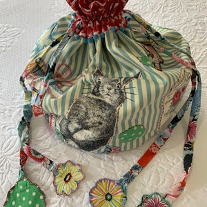 May include: A blue and white striped fabric bag with a red and white floral lining. The bag has a drawstring closure and four fabric tassels with floral designs. The bag features a black and white illustration of a rabbit.