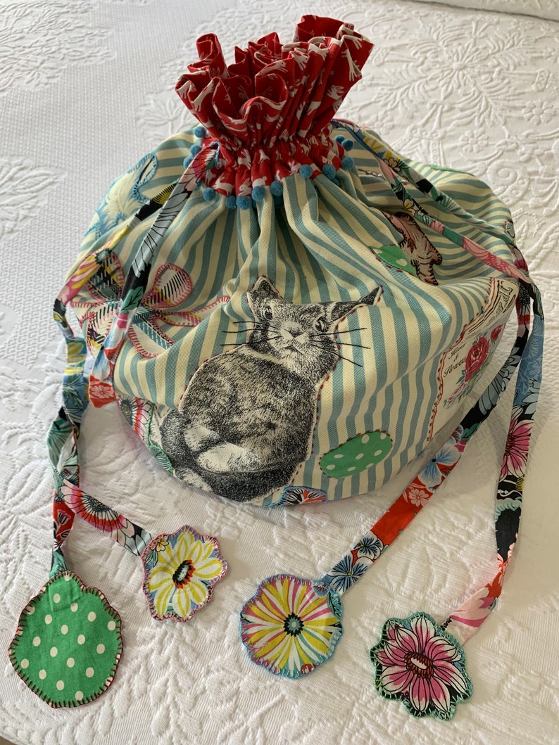 May include: A blue and white striped fabric bag with a red and white floral lining. The bag has a drawstring closure and four fabric tassels with floral designs. The bag features a black and white illustration of a rabbit.