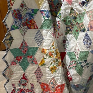 May include: Close-up of a handmade quilt featuring a star pattern. The quilt is made of white fabric with colorful star-shaped patches. The patches are made of various floral and patterned fabrics in red, blue, green, and yellow. The quilt has a striped border.