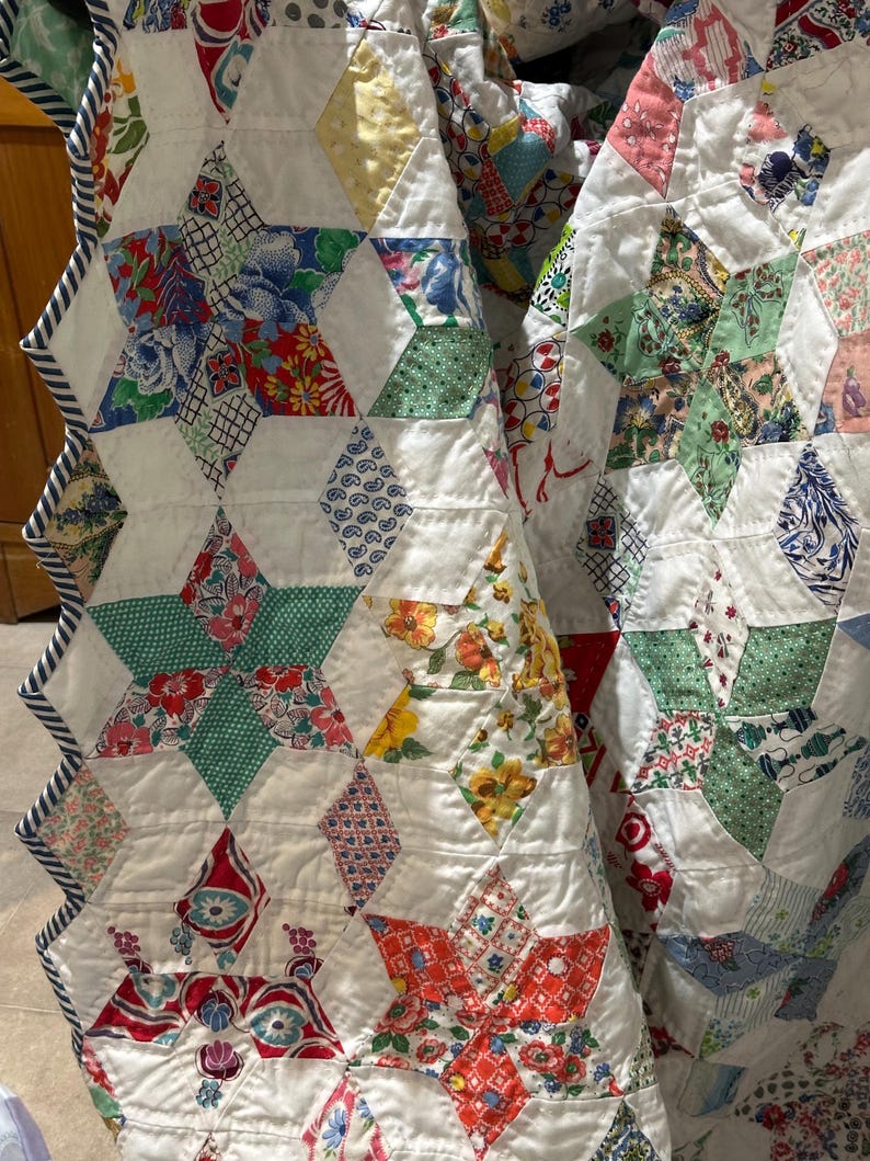 May include: Close-up of a handmade quilt featuring a star pattern. The quilt is made of white fabric with colorful star-shaped patches. The patches are made of various floral and patterned fabrics in red, blue, green, and yellow. The quilt has a striped border.
