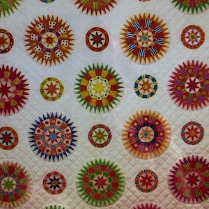 May include: A white quilt with a repeating pattern of colorful, star-shaped designs. The quilt is made up of various shades of red, blue, green, yellow, and orange.