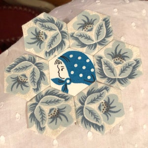 May include: A blue and white fabric hexagon with a floral pattern. The center hexagon features a girl wearing a blue and white polka dot hat.