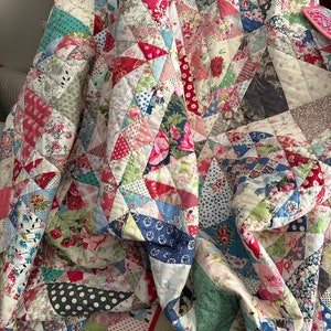 May include: A patchwork quilt made of various floral and geometric fabrics in shades of pink, red, blue, green, and white. The quilt is folded and draped over a chair.