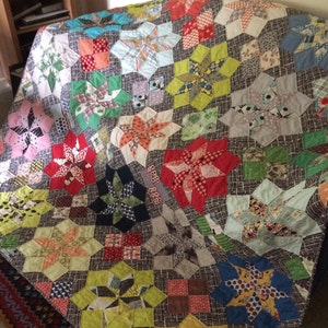 K&ouml;nnte beinhalten: Ein farbenfroher Quilt mit Patchwork-Design, der eine Vielzahl von Stoffen und Farben verwendet, darunter Rot, Blau, Gr&uuml;n, Gelb und Wei&szlig;. Der Quilt besteht aus einer Reihe von sternf&ouml;rmigen Bl&ouml;cken, die jeweils ein anderes Muster aufweisen.