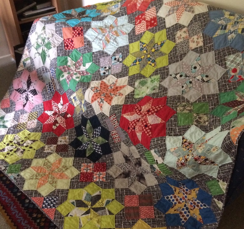 K&ouml;nnte beinhalten: Ein farbenfroher Quilt mit Patchwork-Design, der eine Vielzahl von Stoffen und Farben verwendet, darunter Rot, Blau, Gr&uuml;n, Gelb und Wei&szlig;. Der Quilt besteht aus einer Reihe von sternf&ouml;rmigen Bl&ouml;cken, die jeweils ein anderes Muster aufweisen.