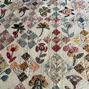 May include: A white quilt with a patchwork design featuring various floral motifs. The quilt is quilted with a wavy pattern and includes a variety of fabrics in shades of pink, red, green, blue, and brown. The quilt is labeled "Calico Garden @msg47".