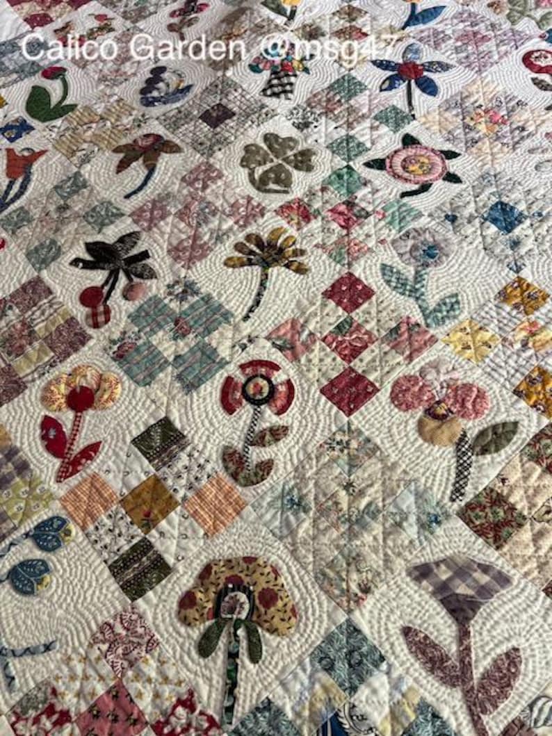 May include: A white quilt with a patchwork design featuring various floral motifs. The quilt is quilted with a wavy pattern and includes a variety of fabrics in shades of pink, red, green, blue, and brown. The quilt is labeled "Calico Garden @msg47".