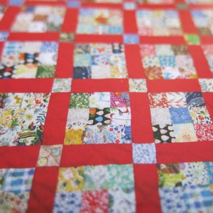 以下が含まれることがあります： パッチワークデザインの手作りキルトのクローズアップ。キルトは、さまざまな色のさまざまな模様の生地の正方形を特徴とし、鮮やかな赤いボーダーで縁取られています。キルトは居心地の良い手作りのアイテムです。